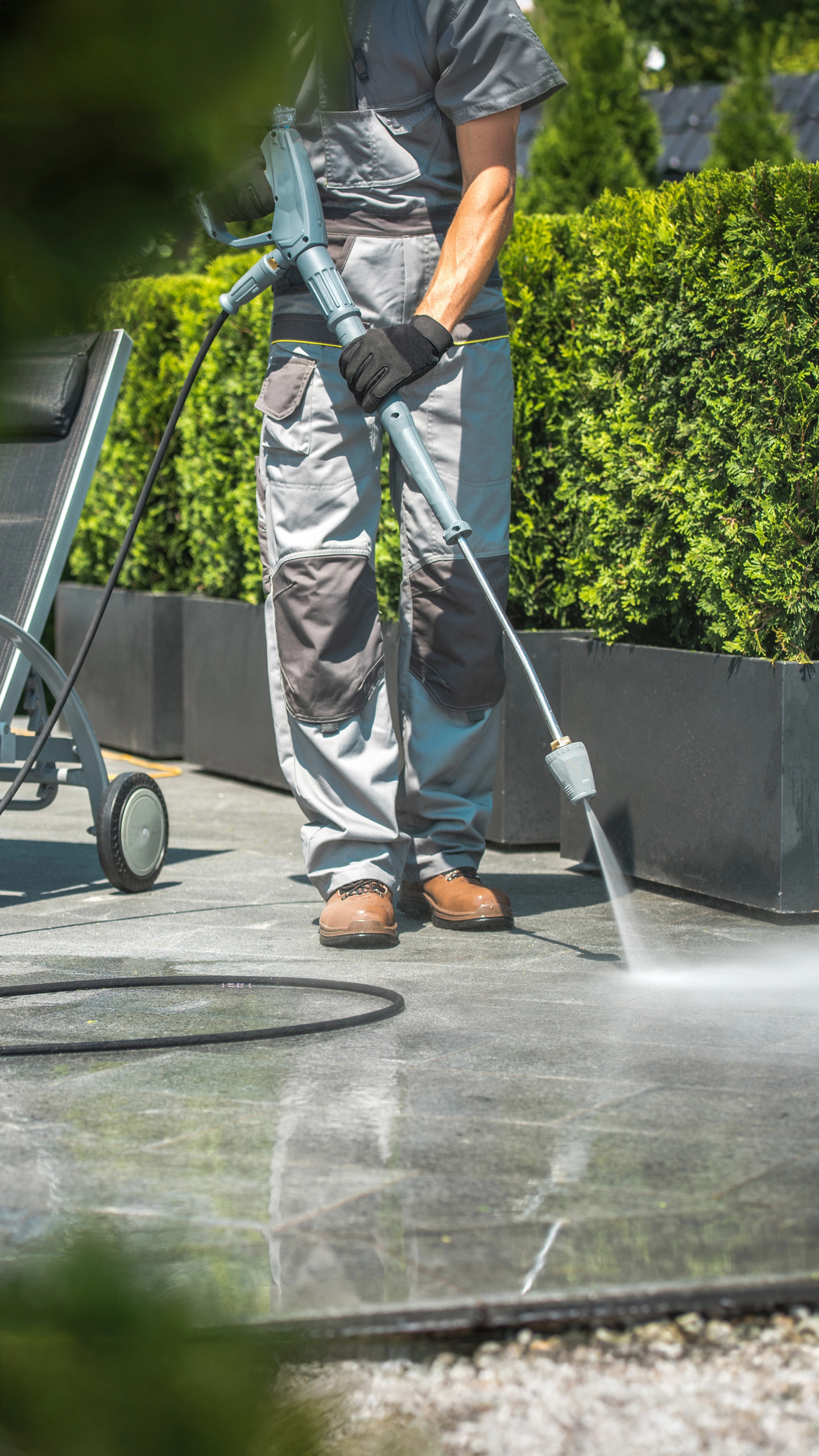 A person wearing gray work attire is using a pressure washer to clean a stone surface in a garden area surrounded by neatly trimmed hedges.